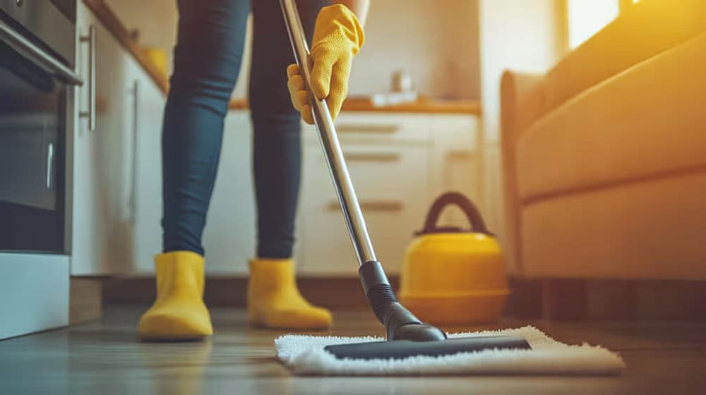 Person in yellow gloves mopping kitchen floor with vacuum cleaner and sunlight streaming in.